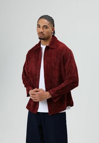 Rode corduroy blouse met knoopsluiting, lange mouwen en een kraag, gecombineerd met een wit T-shirt eronder en donkere spijkerbroek.