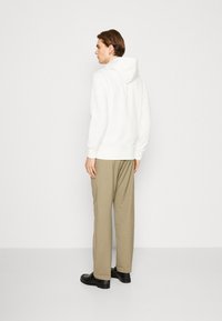 Felpa bianca con cappuccio e polsini a coste, abbinata a pantaloni beige chiaro e scarpe nere, in piedi contro uno sfondo chiaro e uniforme.