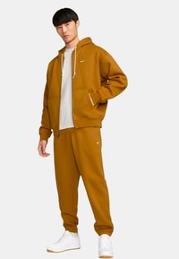 Bruine hoodie met rits en zakken, gecombineerd met bijpassende joggers. Beide gemaakt van soepel materiaal, met een klein wit Nike-logo.