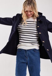Mörkblå kappa med träknappar, har en huva och en öppen framsida, bärs över en vit och mörkblå randig tröja och avslappnade blå jeans.