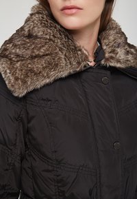 Svart puffjacka med en texturerad brun fuskpälskrage, med tryckknappar och sömdetaljer på kroppen.