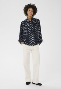 Blouse bleu marine à pois blancs, col noué, manches longues, devant boutonné ; associée à un pantalon large crème et des chaussures noires plates.