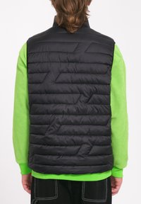 Gilet imbottito nero con motivo a trapunta orizzontale, indossato sopra un maglione verde vivace. Caratterizzato da un colletto alto e senza chiusure visibili.