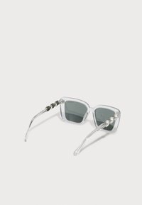 Lunettes de soleil carrées avec monture en plastique transparent, verres gris et accents métalliques décoratifs sur les branches présentant des détails ronds semblables à des bijoux.