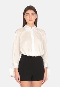 Blusa bianca a pois con colletto strutturato e chiusura a bottoni, caratterizzata da maniche ampie con dettagli ai polsini, abbinata a pantaloni corti neri.