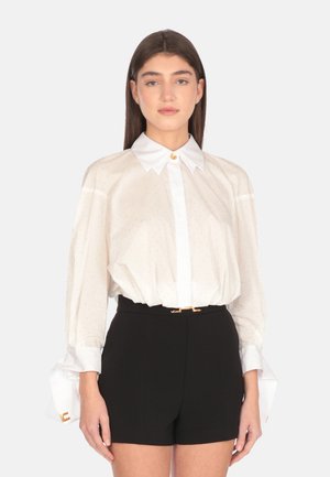 White, polka-dotted blouse with a structured collar and button front, featuring billowy sleeves with cuff accents, paired with black shorts.