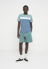 Κοντομάνικη μπλούζα σε μπλε και τιρκουάζ, με λευκή οριζόντια ρίγα και λογότυπο. Συνδυάζεται με τιρκουάζ σορτς και μαύρα αθλητικά παπούτσια.