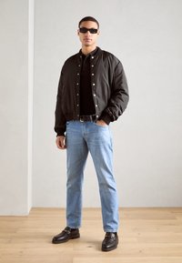 Veste aviateur noire avec fermeture à bouton-pression, associée à un t-shirt noir, un jeans bleu clair et des mocassins sombres sur un fond de sol en bois.