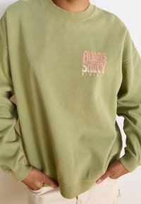 Personne portant un sweat-shirt vert clair avec le texte rose et blanc « Always Salty Roxy », les mains reposant dans les poches d'un pantalon beige.