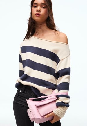 Giovane che indossa un maglione a righe navy e crema con spalle scoperte, che tiene una borsa rosa chiaro su sfondo bianco.