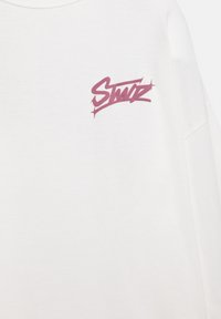 T-shirt blanc à manches courtes en coton avec un logo "Stuz" violet imprimé sur le côté gauche de la poitrine. Texture lisse, coupe décontractée.