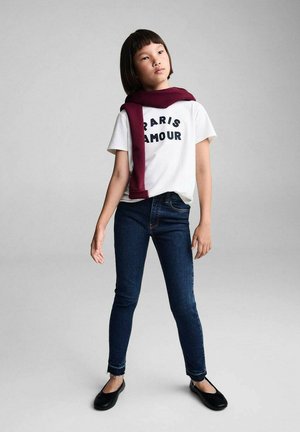 T-shirt blanc avec "PARIS AMOUR" imprimé en noir, associé à un jean skinny bleu foncé. Écharpe bordeaux drapée autour du cou, ballerines noires.