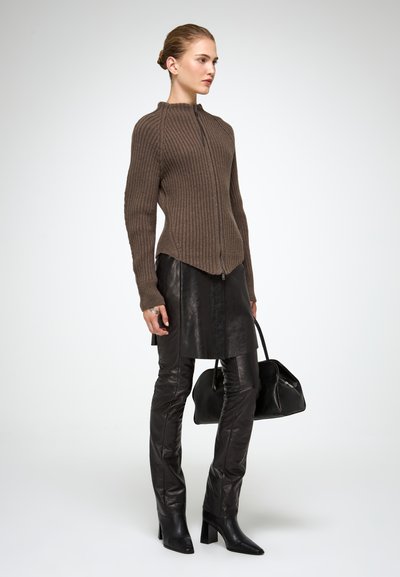 Cardigan marrone a coste con zip abbinato a una gonna di pelle nera e stivali in pelle sopra il ginocchio, mentre tiene una borsa nera.