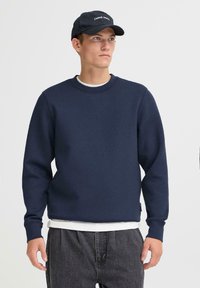Joven vistiendo un suéter azul marino, camiseta blanca debajo, jeans oscuros y una gorra negra con texto, de pie contra un fondo claro liso.