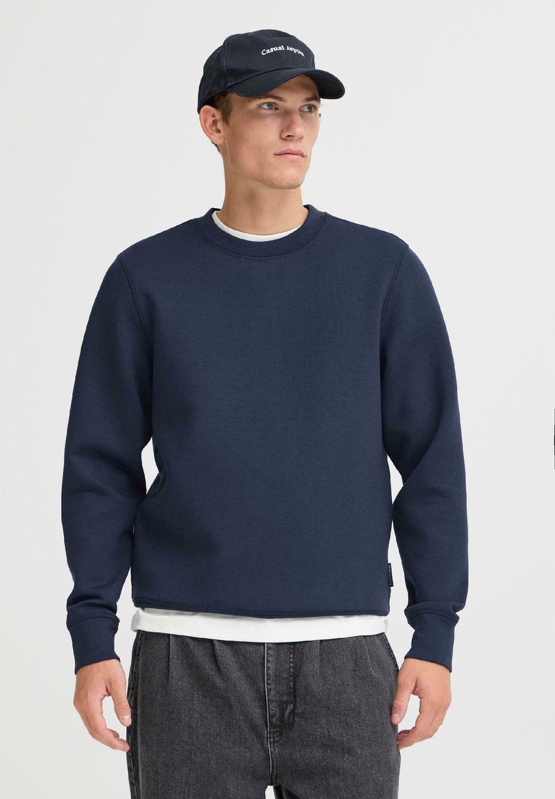 Joven vistiendo un suéter azul marino, camiseta blanca debajo, jeans oscuros y una gorra negra con texto, de pie contra un fondo claro liso.