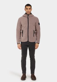 Chaqueta de forro polar en suave marrón claro con cierre frontal de cremallera, capucha y ribetes negros. Cuenta con dos bolsillos laterales y un parche en la manga.