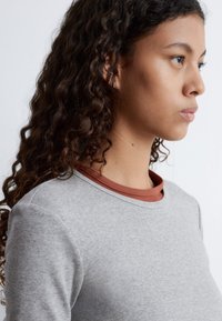 Graues, geripptes Langarmoberteil, das über einem rostfarbenen Rundhalsoberteil getragen wird. Besitzt einen runden Ausschnitt und einen Kontrast in der Textur. Der Hintergrund ist neutral.