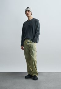 Γκρι μακρυμάνικο πουκάμισο, πράσινο ριγέ φαρδύ παντελόνι, μαύρα αθλητικά παπούτσια και γκρι σκούφος με λογότυπο. Απλή, καθημερινή εμφάνιση πάνω σε ουδέτερο φόντο.