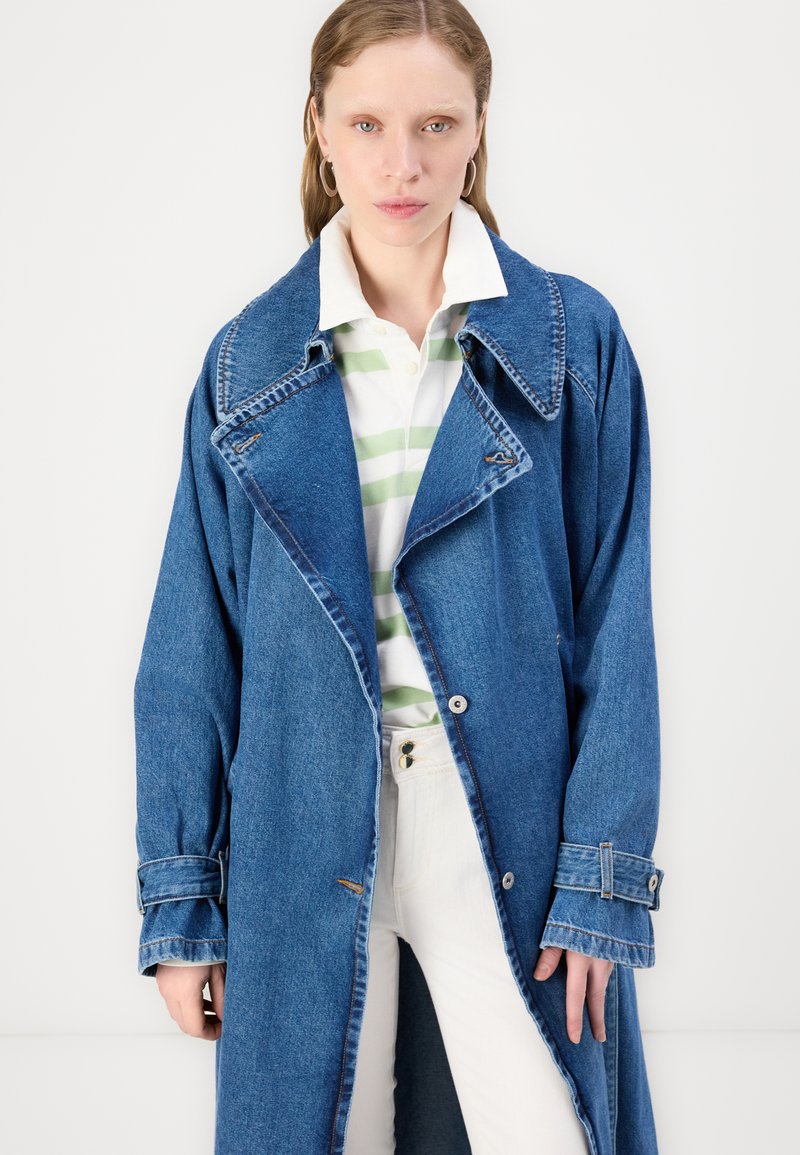 Denim-Trenchcoat, oversized Schnitt, hellblaue Farbe, breite Revers, Knopfverschlüsse und Ärmeldetails. Getragen über einem gestreiften grün-weißen Hemd.