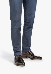 Scarpe in pelle marrone con motivo e finitura lucida, abbinate a pantaloni blu aderenti. Le scarpe presentano una punta rotonda e un laccio frontale.