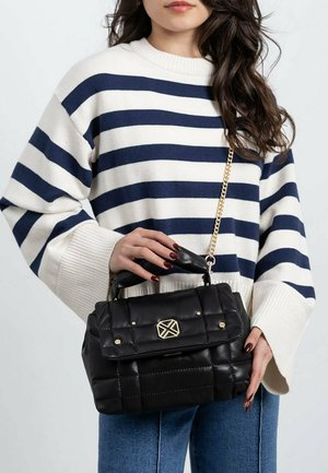 Mujer llevando un suéter a rayas blanco y azul marino, sosteniendo un bolso negro acolchado con cadena de oro y emblema.