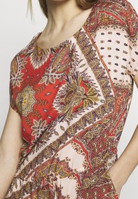 Žena v šatech s krátkým rukávem a elastickým pasem, které mají složitý květinový a geometrický vzor v červené, žluté a béžové barvě.