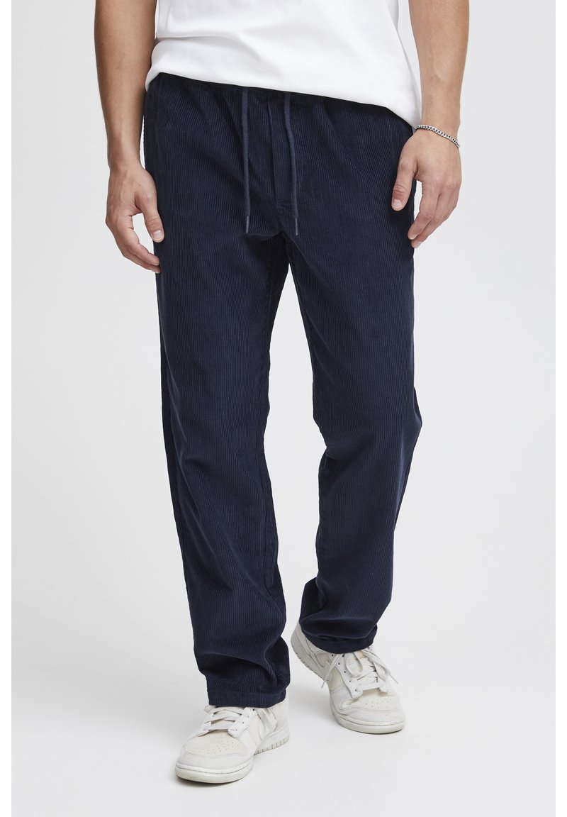 Tmavomodré manšestrové nohavice so šnúrkou v páse, s vertikálnou rebrovanou textúrou a voľným strihom, skombinované s bielymi teniskami.