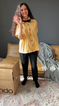 Yellow short-sleeve button-up shirt with sunburst detail, black pants, standing beside a brown cardboard box and a patterned blanket.