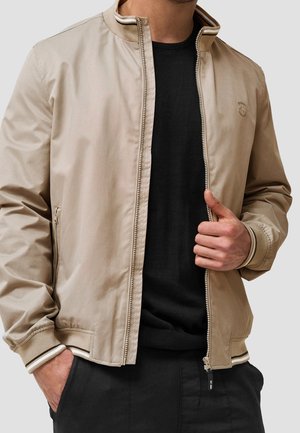 Homme portant une veste beige zippée par-dessus un pull noir, une main tenant la fermeture éclair de la veste et l'autre dans une poche.