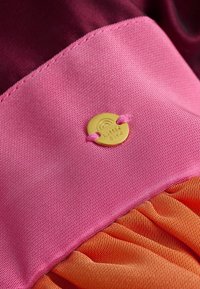 Ein mehrfarbiges Textilmaterial zeigt einen glatten oberen Abschnitt in Pink und einen zerknitterten unteren Abschnitt in Orange, gesichert mit einem runden goldenen Anhänger, der mit "little bird" beschriftet ist.