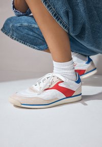 Fehér, piros és kék sportcipők, sima és texturált anyagok kombinációjával, kerek orral, fűzővel és retro dizájnnal, világos háttéren.