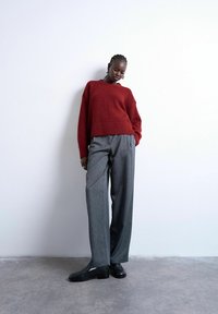 Femme aux cheveux tressés portant un pull rouge, un pantalon large gris et des chaussures noires, se tenant contre un mur blanc sur un sol en béton.