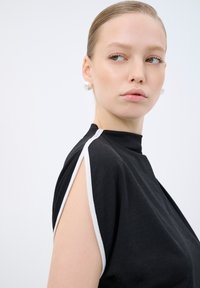 Top nero con scollatura alta, rifiniture bianche sulle spalle e una texture morbida e fluida. La modella indossa grandi orecchini a perla.