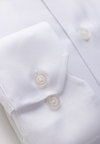 Valge triiksärgi varrukaots kahe läbipaistva nööbiga, mis näitab detailset õmblust ja osa särgi esiplaanist koos nööbiga.