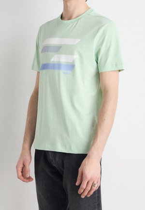 Mężczyzna ubrany w jasnooliwkowy t-shirt z krótkim rękawem z niebieskimi, białymi i szarymi poziomymi paskami, w połączeniu z czarnymi spodniami na gładkim tle.