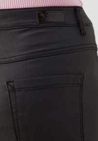 Gros plan d'une poche de pantalon en cuir noir avec détail de couture et étiquette de marque, porté avec un haut en tissu côtelé rose.