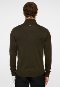 Tumma vihreä neulepaita, jossa on korkea kaulus ja ribatuutiset hihansuut sekä helma. Takana kauluksessa pieni logo ja muotoiltu siluetti.