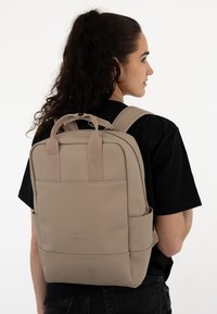 Beige backpack made of smooth material features dual shoulder straps, a top handle, and a front pocket. Minimalist design with subtle branding.