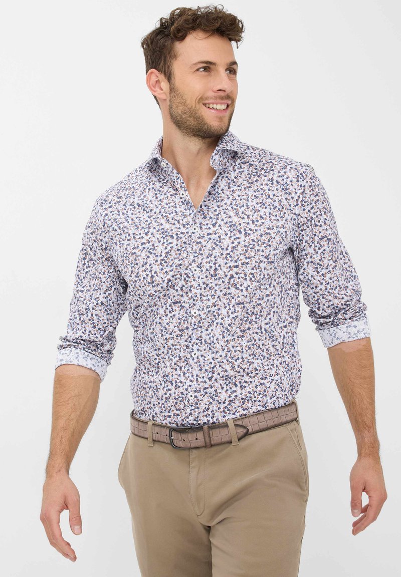 Weißes Hemd mit einem Blumenmuster in Blau und Braun, ausgestattet mit einem Button-Down-Kragen und hochgekrempelten Ärmeln. Kombiniert mit beigen Hosen.