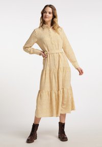 Geel bloemig midi-jurk met een knoopsluiting aan de voorkant, lange mouwen en een ceintuur in de taille. Beschikt over een gelaagd ontwerp en een zachte stoftextuur.