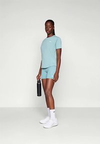 Šviesiai mėlynas sportinis marškinėlių ir šortų rinkinys. Marškinėliai trumpomis rankovėmis su mažu logotipu; modelis laiko juodą vandens butelį ir dėvi baltus sportinius batelius.