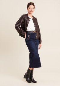 Giacca bomber in pelle marrone, t-shirt bianca, gonna in denim scuro a vita alta con bottoni laterali, stivaletti neri alla caviglia, accenti di gioielli semplici.