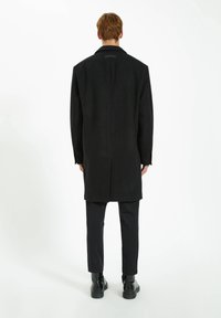Cappotto in lana nera con taglio dritto, colletto largo e maniche lunghe, abbinato a pantaloni neri slim e stivali neri con lacci.