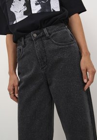 Jeans de mezclilla negros y brillantes con un ajuste relajado. Presentan bolsillos delanteros y costuras visibles. Llevados con una camiseta gráfica negra.