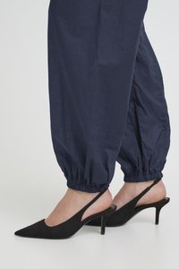 Pėdos avinčios juodus slingback kitten aukštakulnius, derinamus su tamsiai mėlynomis tampriomis kelnėmis su elastinėmis rankogaliais ant baltos fono.