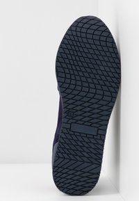 Semelle de chaussure avec une bande de roulement en caoutchouc foncé en motif croisé, taille 38 indiquée, positionnée verticalement contre un mur d'angle blanc.