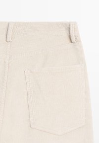 Pantalon en velours côtelé beige avec ceinture, texture à côtes verticales et une seule poche arrière avec un détail de bord surpiqué.