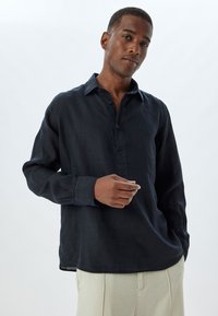 Camisa de lino azul oscuro de Manga Larga con tapeta de botones, textura suave y corte relajado, combinada con pantalones de color claro.