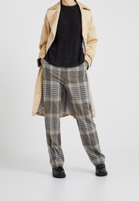 Beige trenchcoat över en svart texturerad tröja och mönstrade rutiga byxor i beige, grå och blå. Svarta skor fulländar looken.