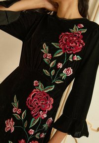 Rochie din catifea neagră cu broderii florale roșii și roz. Are mâneci cu volane și o talie conică, evidențiind modele și texturi intricate.